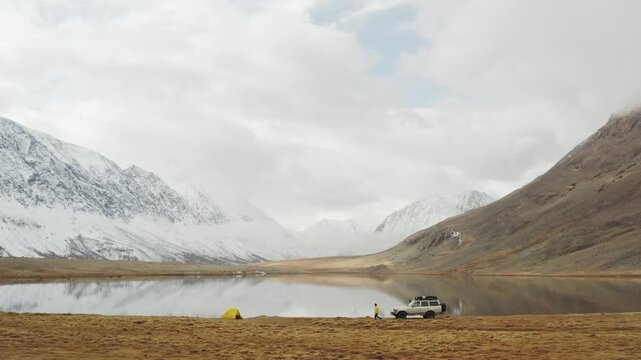Wide landscape with an off road car, yellow tent, and traveler by a calm mountain lake surrounded by snowy peaks and open valley. Remote camping scene combines overland travel, outdoor adventure, and
