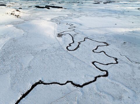 Aerial view of a dark river snaking through a snow-laden landscape, a stark contrast of black against the pristine white, Nasbinals, Occitanie, France.