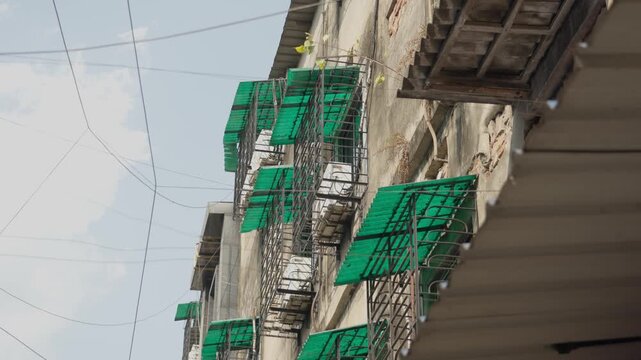 Low angle shot of a weathered residential building facade in India featuring green plastic awnings, AC outdoor unit and tangled power lines against a bright sky.