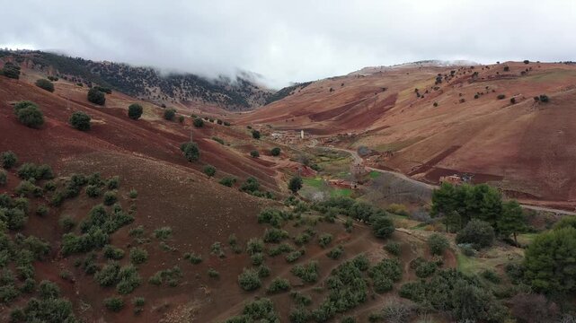 A cinematic 4K aerial wide shot of the Middle Atlas landscape in Morocco, featuring rolling red soil hills, green vegetation, and a distant village tucked into a valley under a blanket of low clouds