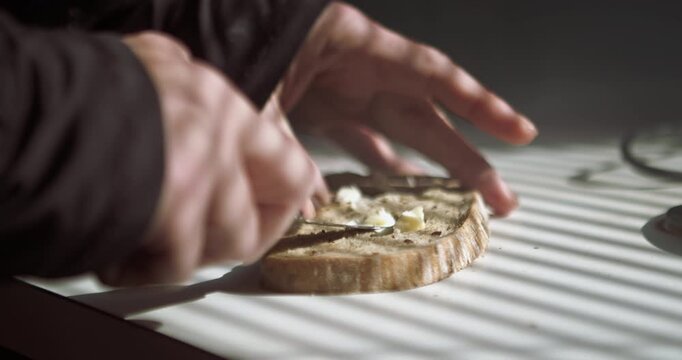 Detailed close-up of hands spreading butter on toast in morning sunblind shadows. Ideal for breakfast commercials, healthy lifestyle vlogs, and domestic routine sequences.