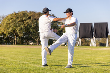 Obraz premium Male cricketers stretching on mown grass, balancing while wearing white kits, sight screens behind