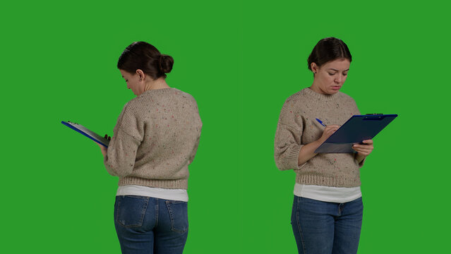 Close up of casual adult analyzing clipboard papers in studio, standing on greenscreen background. Positive woman taking notes on files writing information on documents, working on analysis.