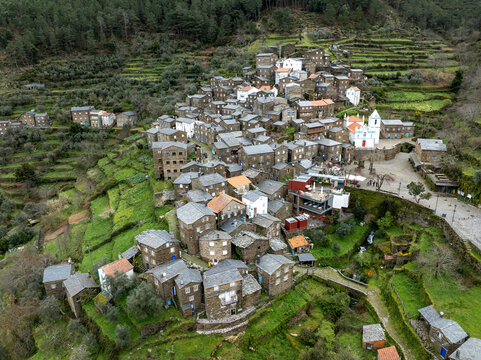 Aerial view of the historical village nestled within terraced hillsides, with a prominent white church standing out against the dark stone buildings, Piodao, Arganil, Portugal.