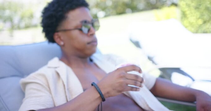 African American man reclining on chair in pink trunks holding glass, sipping to cool down poolside