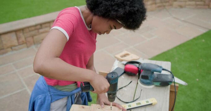 African American teenage female using drill on plank on patio after marking points, checking fit