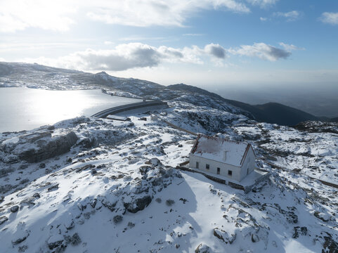 Aerial view of a snow-laden landscape featuring a solitary white building against the backdrop of a serene, partially frozen lake, Lagoa Comprida, Sao Romao, Serra da Estrella, Portugal.