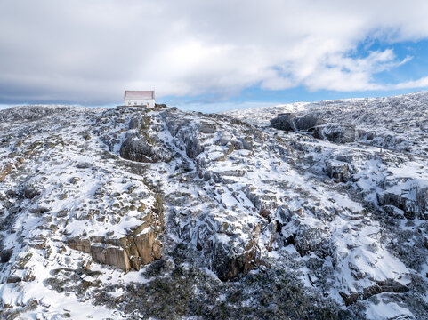 Aerial view of a snow-dusted, rocky peak crowned with a solitary building under a bright sky, Lagoa Comprida, Sao Romao, Serra da Estrella, Portugal.