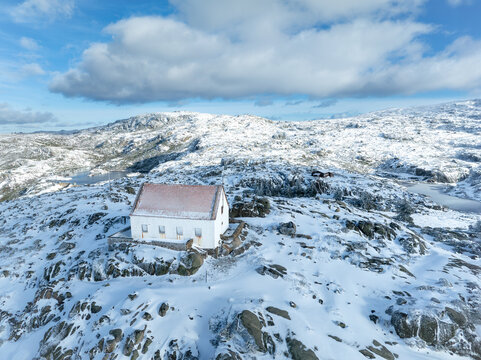 Aerial view of a solitary white house with a red roof sitting atop a snow-covered hill, contrasting against the blue sky, Lagoa Comprida, Sao Romao, Serra da Estrella, Portugal.