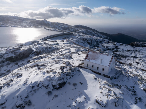 Aerial view of a snow-dusted landscape with a solitary white building overlooking a serene lake reflecting the sky, Lagoa Comprida, Sao Romao, Serra da Estrella, Portugal.
