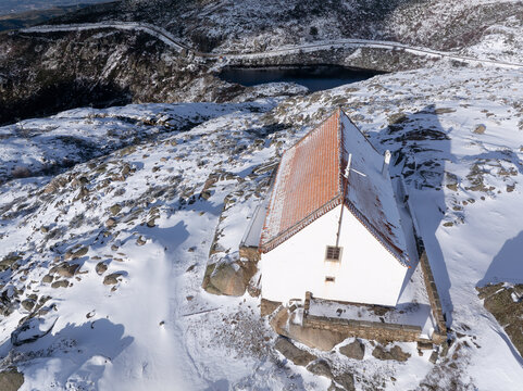 Aerial view of a solitary white building with a red roof amidst a snow-covered landscape near a dark, tranquil lake, Lagoa Comprida, Sao Romao, Serra da Estrella, Portugal.