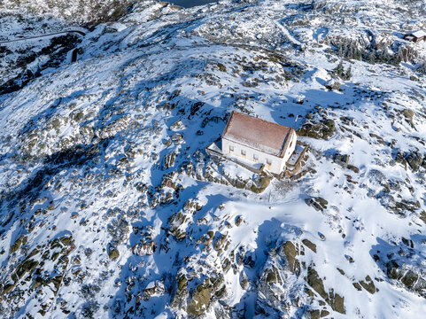 Aerial view of a solitary structure, nestled amidst a snow-dusted, rocky landscape, with the tranquil lake shimmering in the distance, Lagoa Comprida, Sao Romao, Serra da Estrella, Portugal.