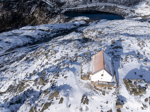 Aerial view of a snow-dusted landscape with a small white building and a dark lake in the distance, Lagoa Comprida, Sao Romao, Serra da Estrella, Portugal.