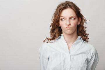 Obraz premium Young woman with curly hair in white shirt looks sideways with skeptical expression on light background. Concept of doubt, suspicion, and uncertain emotions.