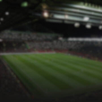 An abstract, out-of-focus view of a professional football stadium during a night match. The image shows a lush green pitch under intense stadium lights with a blurred audience in the stands. This boke