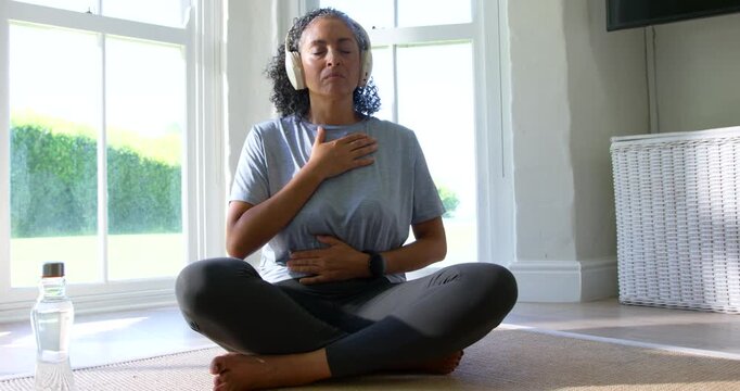 African American woman wearing headphones, following guided audio breathing on mat by bay window