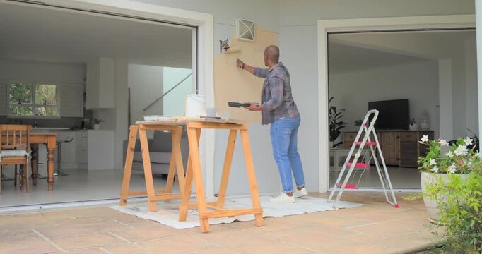 African American man touching-up wall patch on porch using roller; phone ringing, answering smiling
