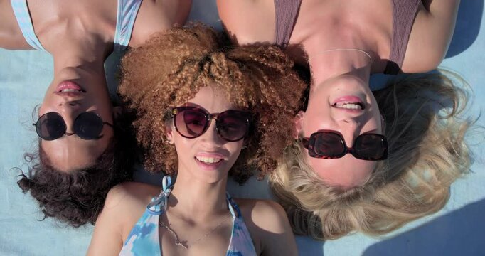 3 female friends lying on towel camera filming above center woman speaking to camera lifting shades