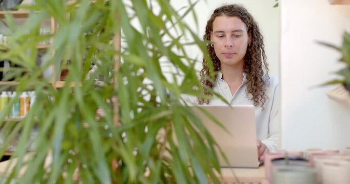 Camera pulling back plant revealing African American nonbinary person using laptop at desk for work