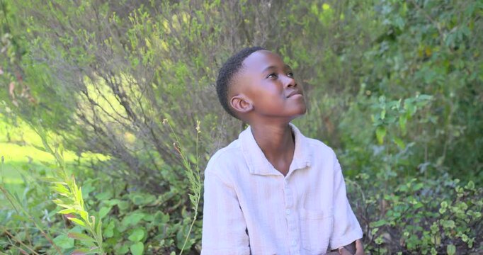 African teenage male sitting in shrubs by stem in park seeing motion up uncrossing turning smiling