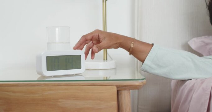 Alarm ringing, woman in green sleepwear pressing clock beside bed, by glass, snoozing, copy space
