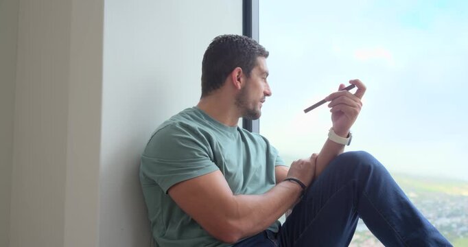 Adult man sitting on window ledge holding smartphone near mouth, glancing outside, tapping screen