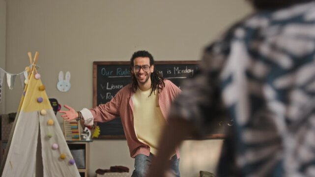 Handheld tilt down shot of cheerful young biracial male kindergarten teacher greeting happy Caucasian children running towards him and hugging together in cozy modern classroom