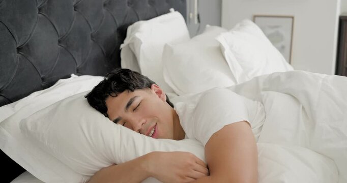 Asian man lying in bed in white T-shirt, hugging white pillow, smiling then fading into sleep