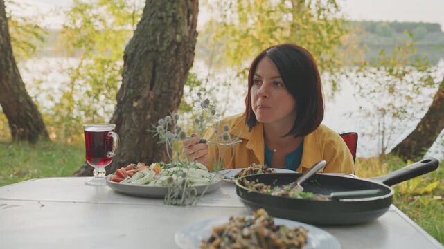Caucasian woman camping by lake camper chef testing recipe with skillet, stirring herbs and plating salad, golden sunset light over water, contemplative tasting, culinary experiment in nature, rustic