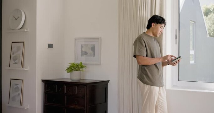 Asian adult man tapping and scrolling tablet while leaning on curtain at bedroom window, copy space