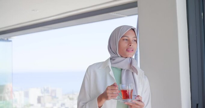 African American woman in gray hijab lifting cup, saucer, sipping at highrise window admiring view