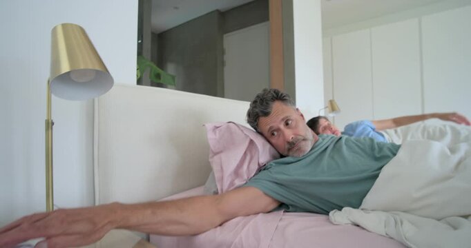 Couple in bed waking to alarm ringing, man reaching digital clock in teal T-shirt, woman cuddling