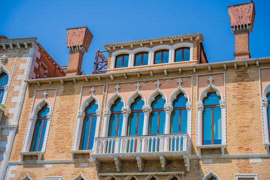 Venice, Italy - 29 March 2026: View of an ornate building facade with arched windows reflecting the azure sky, framed by salmon and brick hues.
