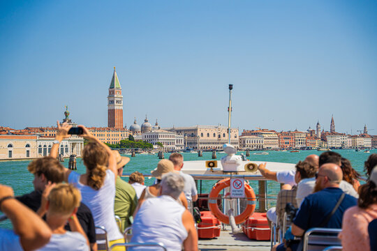 Venice, Italy - 29 March 2026: View of tourists gazing toward St. Mark's Campanile and the Doge's Palace under a vast, cloudless sky, with the serene water reflecting the city's warm hues.