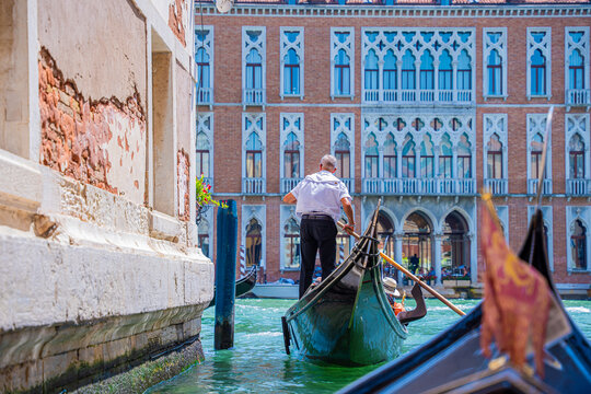 Venice, Italy - 29 March 2026: View of a gondolier navigating the vibrant green waters of a canal, framed by weathered walls and the ornate facade of a building.