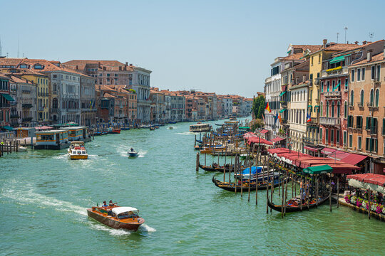 Venice, Italy - 29 March 2026: View of the Grand Canal shimmering under a bright sky, with gondolas and boats navigating past historic buildings and vibrant waterfront cafes.
