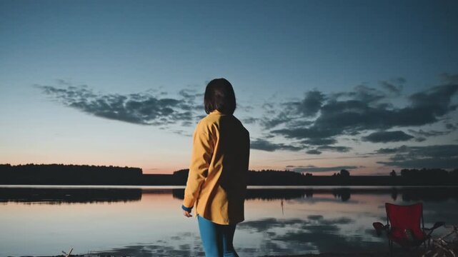 Woman at lake during sunset camping, yellow jacket silhouette on shore facing calm water, red chair and fishing rod nearby, peaceful twilight sky with cloud reflection and soft breeze, contemplative