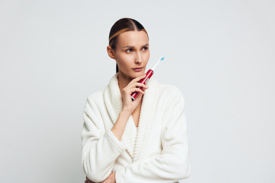 A young woman in a white bathrobe holds a red electric toothbrush near her face, looking thoughtfully to the side against a plain white background. Concept for dental care and morning routine.