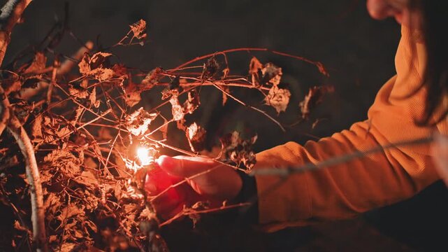 Glowing flame woman lighting dry twigs at lakeside campsite, flame grows among branches, warm orange illumination and smoke, closeup hand holding match, cozy nighttime comfort and satisfaction