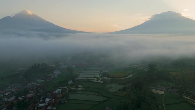 A cinematic sunrise scene over Mount Sindoro and Mount Sumbing in Central Java, Indonesia, with thick morning mist covering the valley and rice fields below.