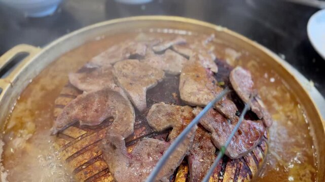 Stainless steel tongs flipping pork liver on a hot brass barbecue pan. Close-up grilling action with smoke and sizzling texture, showing cooking process in restaurant style.