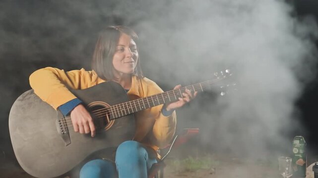 Evening filled with cheerful tunes and bright flames. Night echoes with joyful sounds as woman serenades around campfire. Laughter and harmonious singing animate evening by glowing and crackling fire