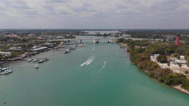 Drone flying west past the Jupiter Lighthouse over the Jupiter Inlet toward the Carlin White Bridge and US 1 with traffic crossing the bridge and many boats on the water and DuBois Park to the south.