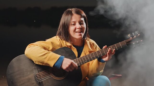 Woman performs music outdoors tonight. Solitary woman music artist performs under stars beside outdoor fire. Single female vocalist entertains outdoors beside crackling campfire beneath night sky