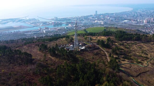 Aerial View of TV Radio 5G Transmitter Tower High Above Swansea City with Marina in Distance. Communication Infrastructure for Smart Phones and Connectivity.
