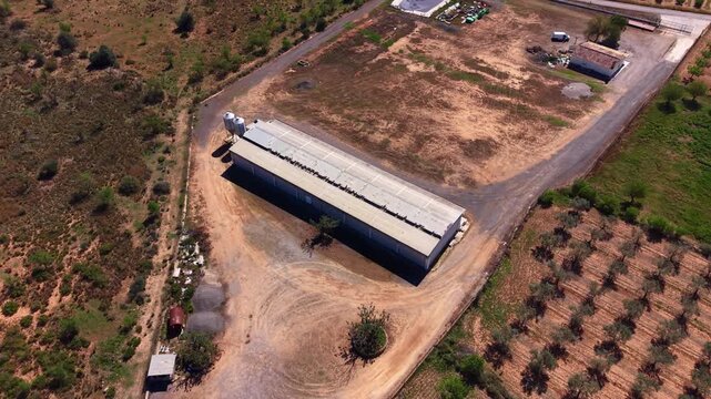 Overhead view of a long rectangular farm shed surrounded by dry dirt roads, scrubland, storage tanks, and rural outbuildings in a semi-arid landscape