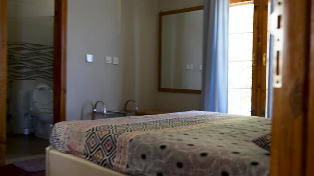 Neatly made bed beside a window with curtains and mirror reflecting soft daylight in a residential room