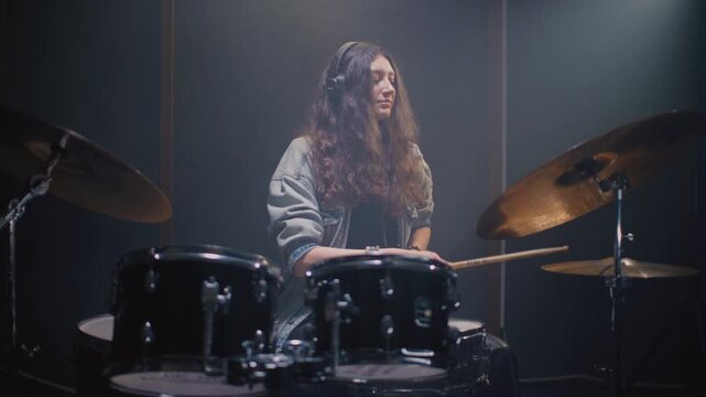 Beautiful drummer girl with long hair is tapping rhythm and drum part in rehearsal room. Young female with headphones performs solo on stage, kit. Sound checking equipment before a rock concert