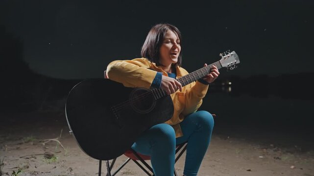 Energetic outdoor party with singing woman. Lively campfire scene with smiling Asian guitarist performing. Cheerful female musician entertaining friends around warming fire under night sky