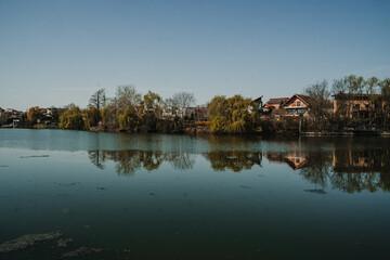 Fototapeta premium Peaceful lake landscape with reflections of houses and autumn trees in calm water. Quiet suburban scenery with natural reflections and soft light.